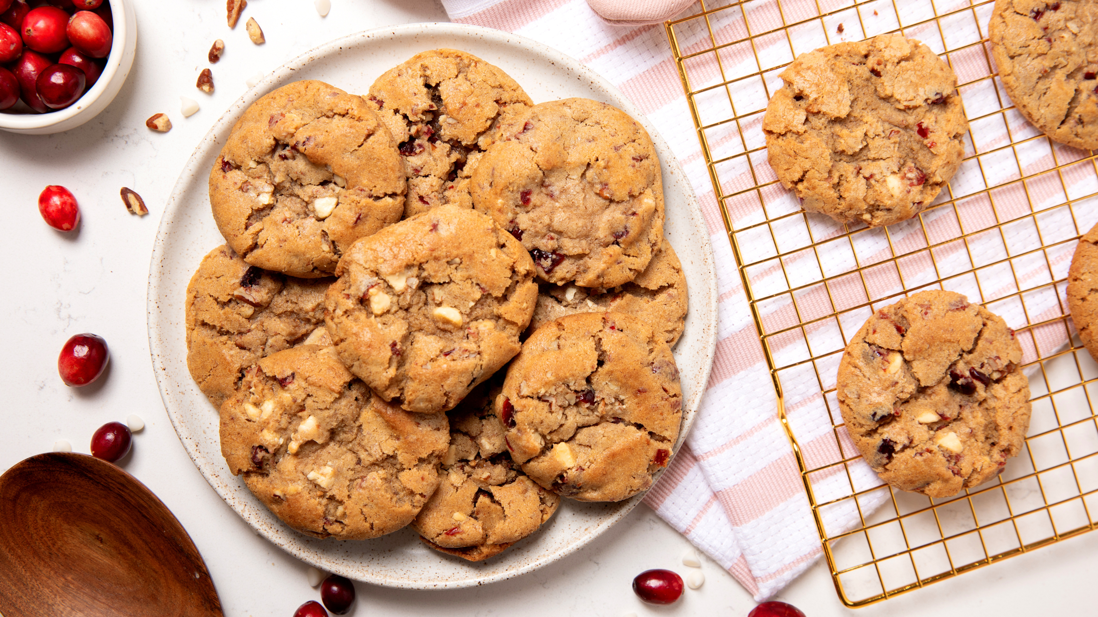 Freshly baked cranberry and pecan cookies on a plate and cooling rack, surrounded by scattered cranberries and pecan nut shavings.